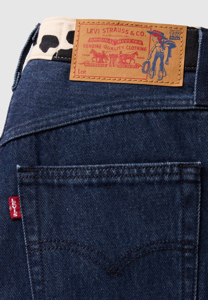 Denim jeans med en mørkeblå vask, læderlabel med rød trykt grafik og kontrasterende sort-hvid accent ved taljen.