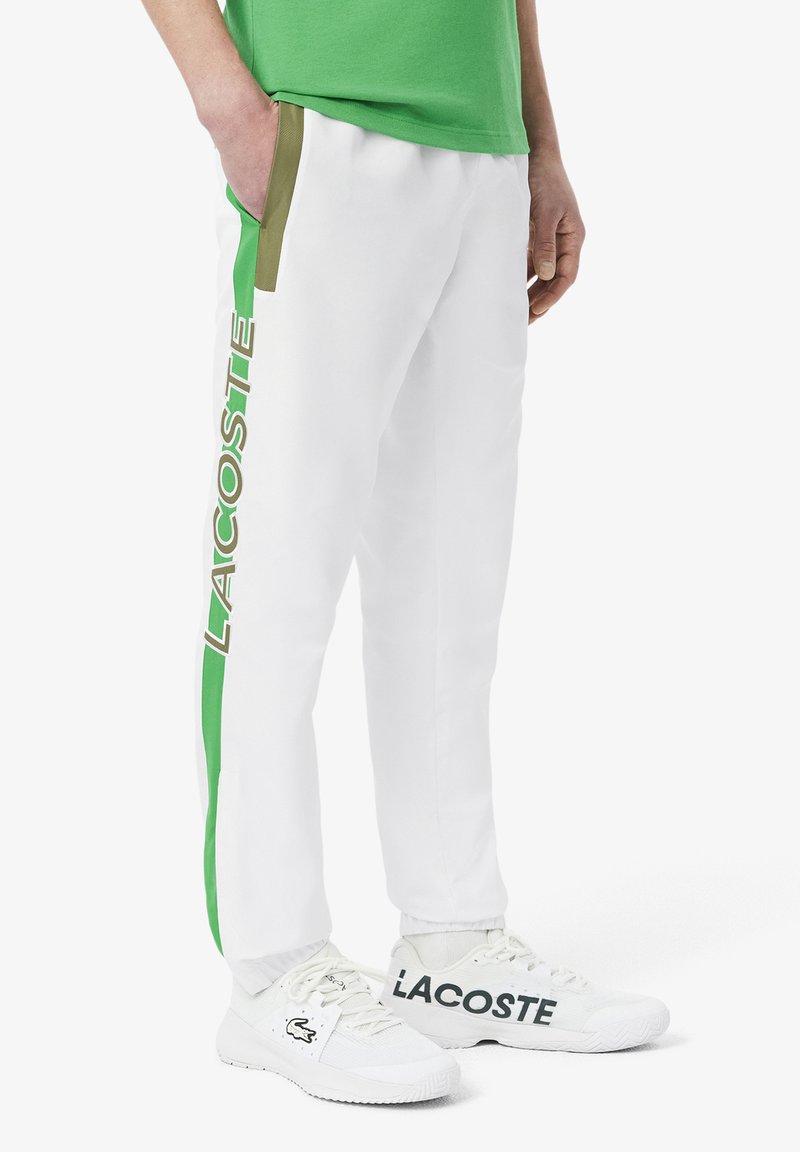 Weiße Jogginghose mit grünen Akzenten und "LACOSTE"-Schrift entlang der Seite, ausgestattet mit einem elastischen Bund und Taschen. Kombiniert mit weißen Sneakers.
