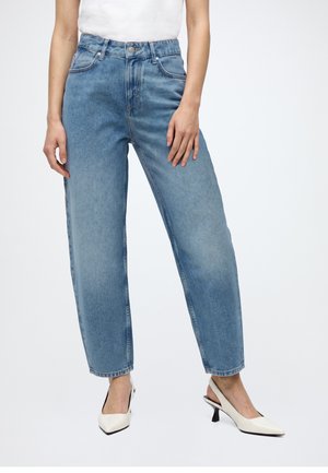 Femme portant un jean bleu clair droit, un haut blanc sans manches et des talons slingback blancs à bout pointu, debout sur un fond blanc.