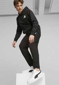 Hoodie de algodão preto com bolso canguru e logo, combinado com calças de fato pretas. O modelo usa ténis brancos com detalhes em azul-marinho, em pé sobre um pedastal.
