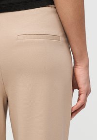 Κοντινό πλάνο της λεκάνης και του χεριού ενός ατόμου που φοράει μπεζ παντελόνι με τσέπη welt και μαύρο πάνω μέρος.