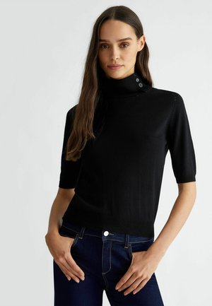 Maglione corto a maniche corte di colore nero con una forma aderente, texture liscia e dettagli con bottoni sul colletto. Indossato con jeans di denim scuro.