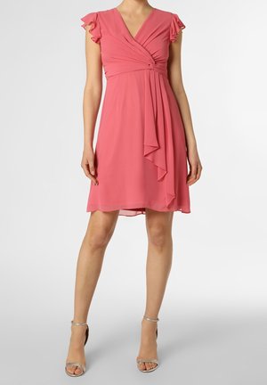 Kobieta w różowej sukience kopertowej do kolan z falbaniastymi rękawami i srebrnymi sandałami na obcasie, stojąca na jednolitym tle.