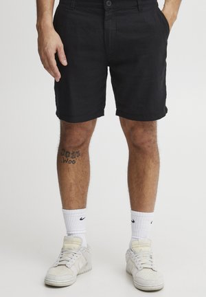 Zwarte linnen shorts met een knoopsluiting, zijzakken en opgerolde randen. Draagt witte sokken en sneakers met een gestructureerde afwerking.