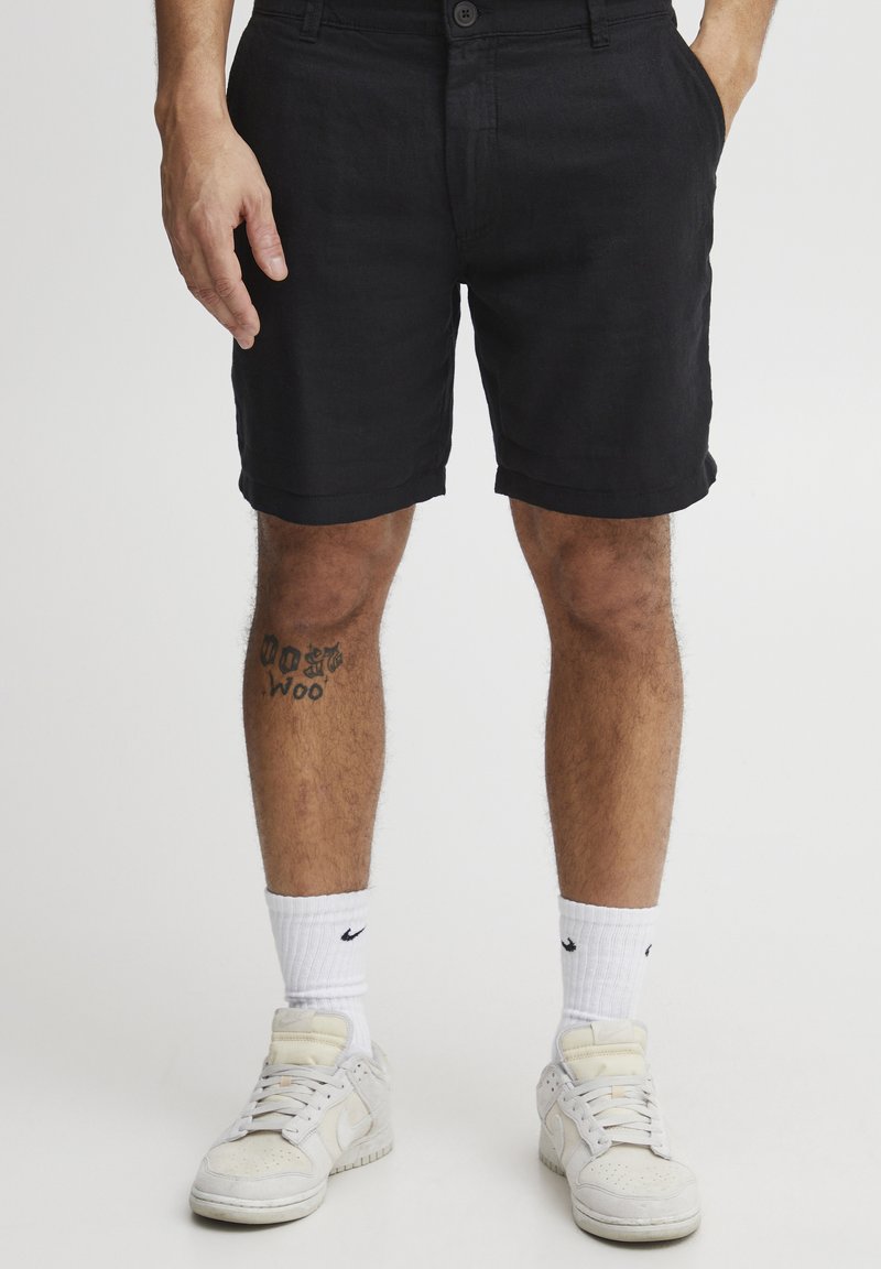 Svarte linshorts med knappelukking, sidelommer og rullet kant. Brukt med hvite sokker og joggesko med tekstur.
