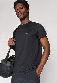 Μαύρη αθλητική μπλούζα με κοντά μανίκια, με υφή υφάσματος και μικρό λογότυπο στο στήθος. Φοράει με σκούρες βερμούδες και τσάντα ώμου.