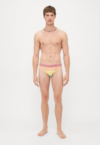 Junger Mann, der barfuß vor einem weißen Hintergrund steht und bunte Dsquared2-Unterwäsche sowie eine Perlenkette trägt.