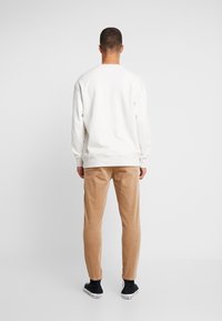 Kremowa bluza z opuszczonymi ramionami, zestawiona z beżowymi spodniami sztruksowymi. Czarne trampki dopełniają strój. Gładkie tło.