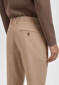 Man wearing beige tailored trousers and a brown long-sleeve top, shown from the back waist to mid-thigh.
