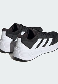 Mustat urheilukengät, joissa on hengittävä verkko-osa, valkoiset yksityiskohdat ja kolme valkoista raitaa sivussa. Varustettu pehmeällä pohjalla ja teksturoidulla kantapäällä.