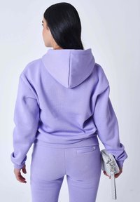 Felpa con cappuccio lilla e pantaloni della tuta abbinati in un tessuto morbido e strutturato, con tasca a marsupio e una piccola tasca posteriore. Viene tenuta in mano una pochette bianca con motivo.