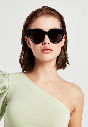 Black oversized cat-eye sunglasses with dark lenses, featuring a sleek, smooth design and bold frame. Complemented by a light green top.