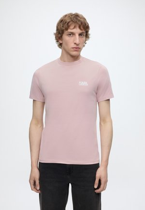 Jeune homme aux cheveux bouclés portant un t-shirt rose pâle Karl Lagerfeld et un pantalon noir, debout devant un fond blanc.