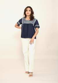 Haut tissé bleu marine avec des motifs brodés blancs, coupe décontractée ; associé à un pantalon droit blanc et des sandales légères à lanières.