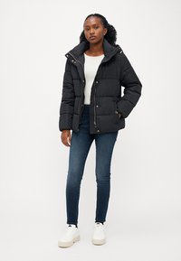 Svart pufferjakke, quiltet design, med hette og glidelås. Kombinert med blå jeans og hvite sneakers. Enkel og funksjonell stil.