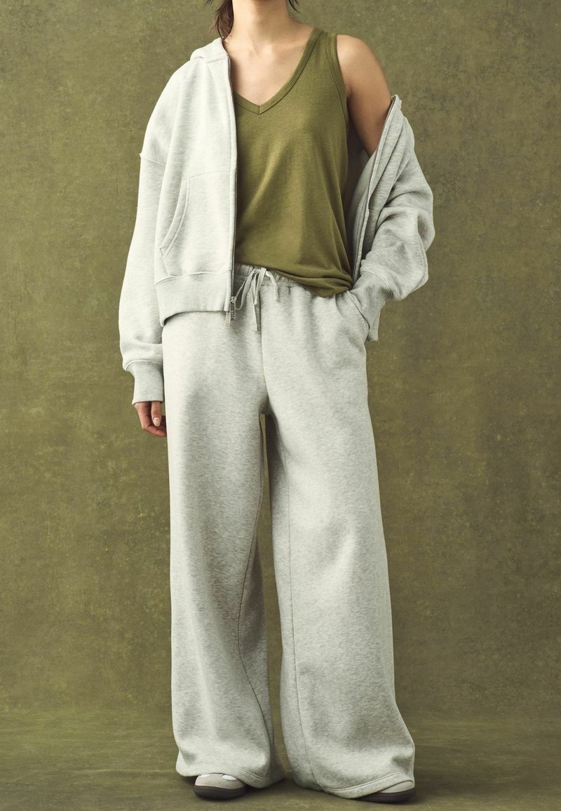 Personne portant un pantalon de survêtement gris clair à jambes larges, un sweat à capuche zippé assorti tombant sur les épaules, un haut sans manches vert olive et des baskets.