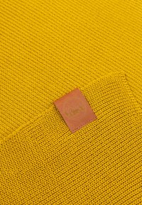 Dzianinowy żółty materiał z teksturowanym wzorem, z małą brązową skórzaną metką z wytłoczonym logo.