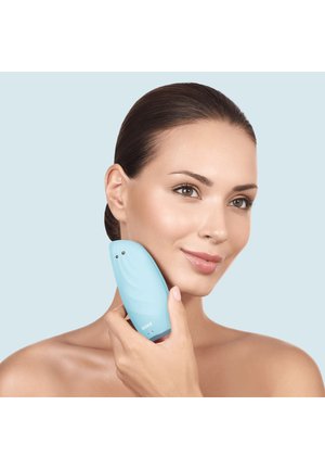 Femme à la peau claire tenant une brosse nettoyante bleue pour le visage contre sa joue sur un fond bleu clair neutre.