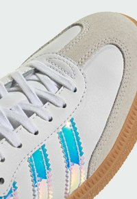 Baskets blanches avec embout en daim beige, lacets blancs, semelle gomme et bandes bleues iridescentes sur le côté.