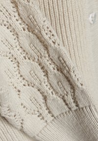 Stickad tröja i krämfärgad med ett mönster av kabelstickning på ärmarna, med en ribbad nederdel och tre knappar på framsidan. Texturerad yta.