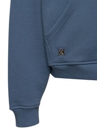 Plava majica dugih rukava od teksturiranog materijala, s džepom, rebrastim manžetnama i prepoznatljivim crnim grbom na donjoj strani.