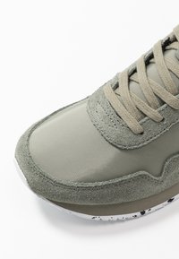 Basket de sport avec une tige vert olive alliant matériaux synthétiques lisses et suède texturé. Fermeture à lacets avec des lacets beiges plats.