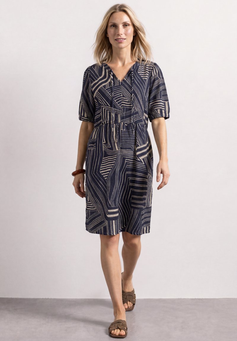 Femme marchant, portant une robe noire mi-longue avec des lignes géométriques beige, un bracelet marron et des sandales marron à bout ouvert sur un sol gris et un mur blanc.