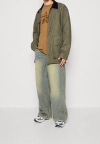 Olive green lightweight jacket with black collar, brown graphic t-shirt, and baggy light blue jeans. Silver sneakers complete the outfit.