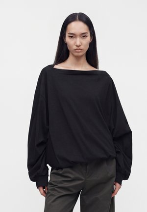 Jeune femme aux longs cheveux noirs porte un haut noir à manches longues surdimensionné et un pantalon gris foncé, debout devant un fond clair uni.