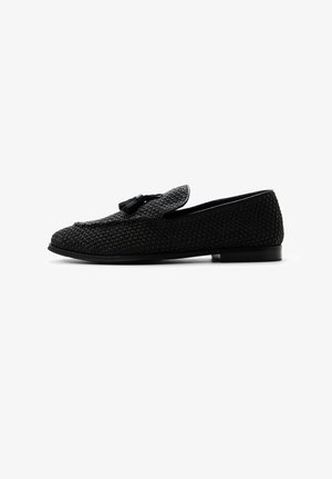 Zwarte geweven loafer met tassel-detail bovenop, lage hak en ronde neus, zijaanzicht op een witte achtergrond.