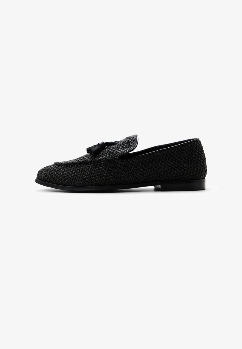 Mocasín tejido negro con detalle de borlas en la parte superior, tacón bajo y punta redondeada, mostrado de lado sobre un fondo blanco.