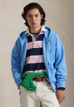 Lyseblå hættetrøje med lynlås over polotrøje med mørkeblå og pink striber, grøn strikket sweater bundet om livet og cremefarvede cargobukser med synligt hardware.