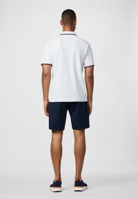 Muž v bielej polokošeli s krátkym rukávom, námorníckych šortkách a námorníckych teniskách stojí chrbtom k prázdnemu pozadiu.