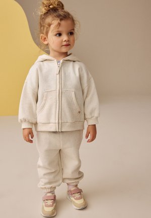 Copil mic cu părul creț prins într-un coc înalt, purtând o geacă cu glugă crem prevăzută cu fermoar, pantaloni asortați și adidași cu accente roz, stând în interior.