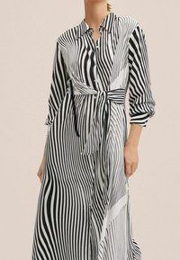 Femme portant une robe boutonnée à rayures verticales noires et blanches avec des manches longues et une ceinture nouée à la taille.