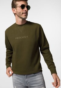 Oliwkowy sweter z okrągłym dekoltem i długimi rękawami, z subtelnym haftowanym logo na klatce piersiowej. Noszony z wyblakłymi szarymi dżinsami.