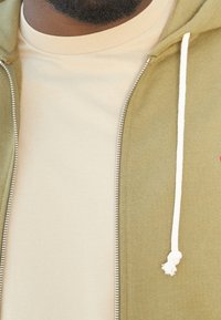 Felpa con cappuccio verde oliva con cerniera e cordoncini bianchi sopra una t-shirt beige chiaro. Il tessuto sembra morbido con una texture liscia.