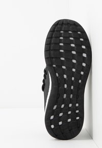 Suela de zapatilla negra con surcos estampados y acentos blancos, posicionada verticalmente contra una pared blanca en la esquina.