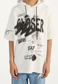 Vit oversized hoodie med grafisk svart text och illustrationer, snörning i huven, korta ärmar, framficka med blandade textelement.