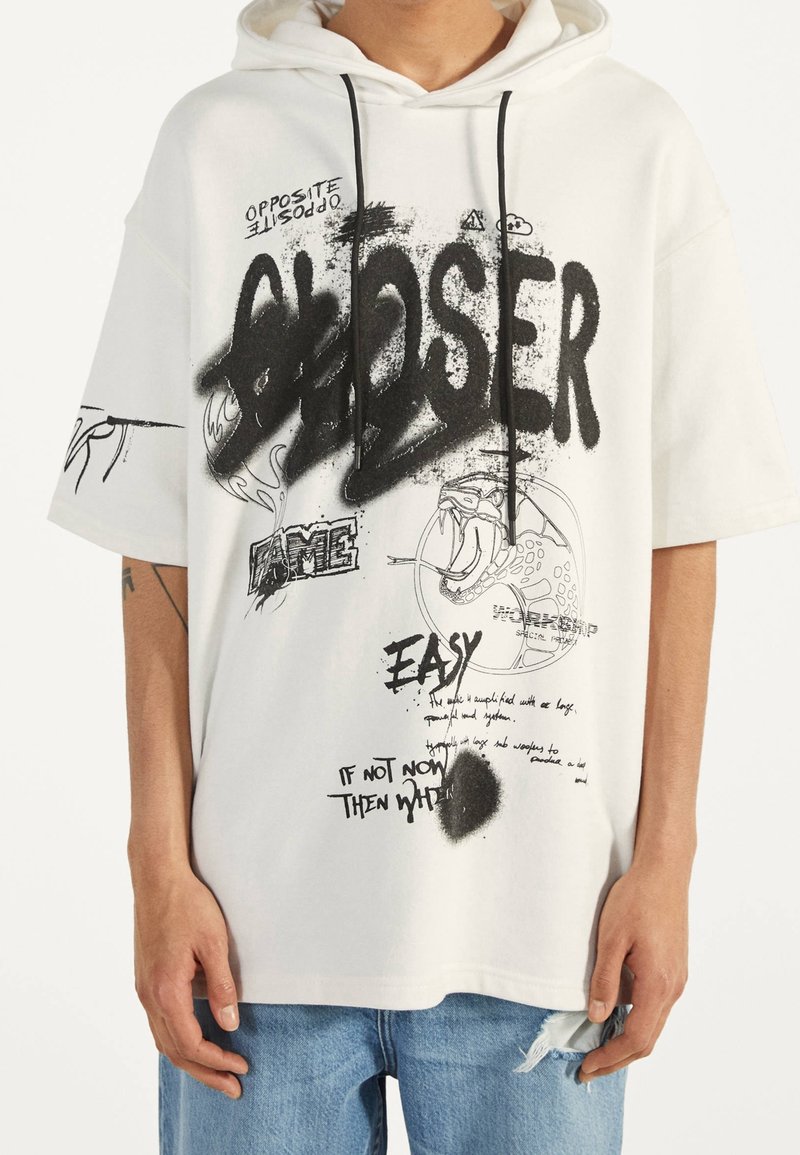 Vit oversized hoodie med grafisk svart text och illustrationer, snörning i huven, korta ärmar, framficka med blandade textelement.