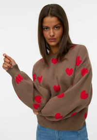 Femme aux longs cheveux noirs portant un pull marron avec des cœurs rouges et un jean bleu, posant devant un fond blanc uni.