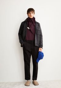 Herenoutfit met een donkergrijze, gestructureerde wollen jas met knopen, een bordeauxrode top, zwarte broek, beige schoenen en een blauwe pet.