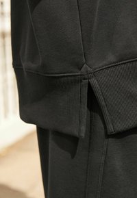 Černá mikina s žebrovaným lemem a bočními rozparky. Měkká textura, design s kulatým výstřihem a viditelné švy zvýrazňují její strukturu.