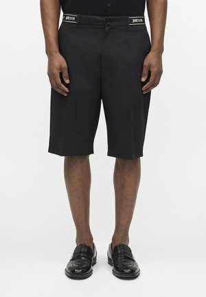 Man draagt zwarte op maat gemaakte knielange shorts met "Just Cavalli" labels nabij de zakken en zwarte leren loafers.