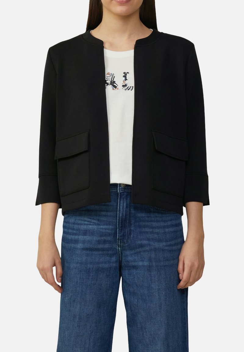 Zwarte cropped jasje met een ronde halslijn, twee voorkruiken, en driekwartmouwen, gedragen over een witte shirt met een grafisch ontwerp.