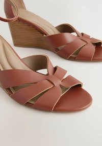 Sandalias de cuña de cuero marrón con un diseño entrecruzado, punta abierta y tacón de madera texturizada. Cuenta con un forro interior suave.