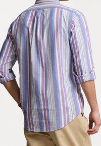 Camisa a rayas con líneas verticales azules, rojas y blancas, mangas largas arremangadas en los puños, cuello con botones y un dobladillo curvado.