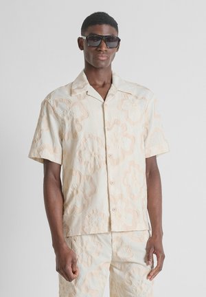 Man die een crème kleurig, korte mouwen, knoophemd en bijpassende broek met grote bloemrijke borduursels draagt, geaccentueerd met donkere zonnebril.