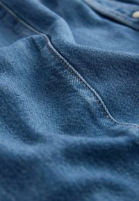 Prim-plan cu material din denim albastru, cu cusături vizibile și detalii texturate ale țesăturii.