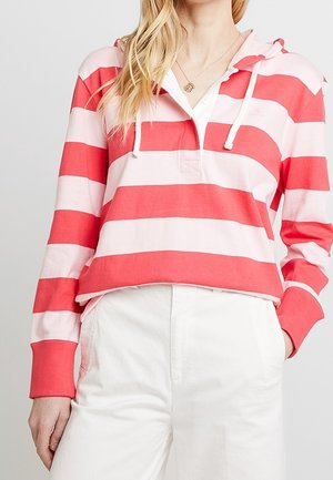 Mujer con sudadera con capucha de rayas anchas en rosa y rojo, con cordones blancos, y pantalones blancos de talle alto, frente a un fondo liso.